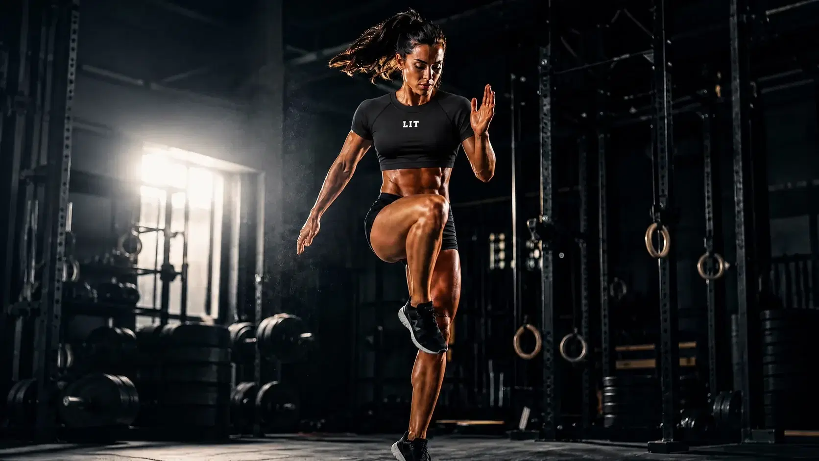An athlete in the black LIT Fitted Crop Tee driving a knee high mid-stride in a dark gym, edge-lit by a single hard window light behind her.