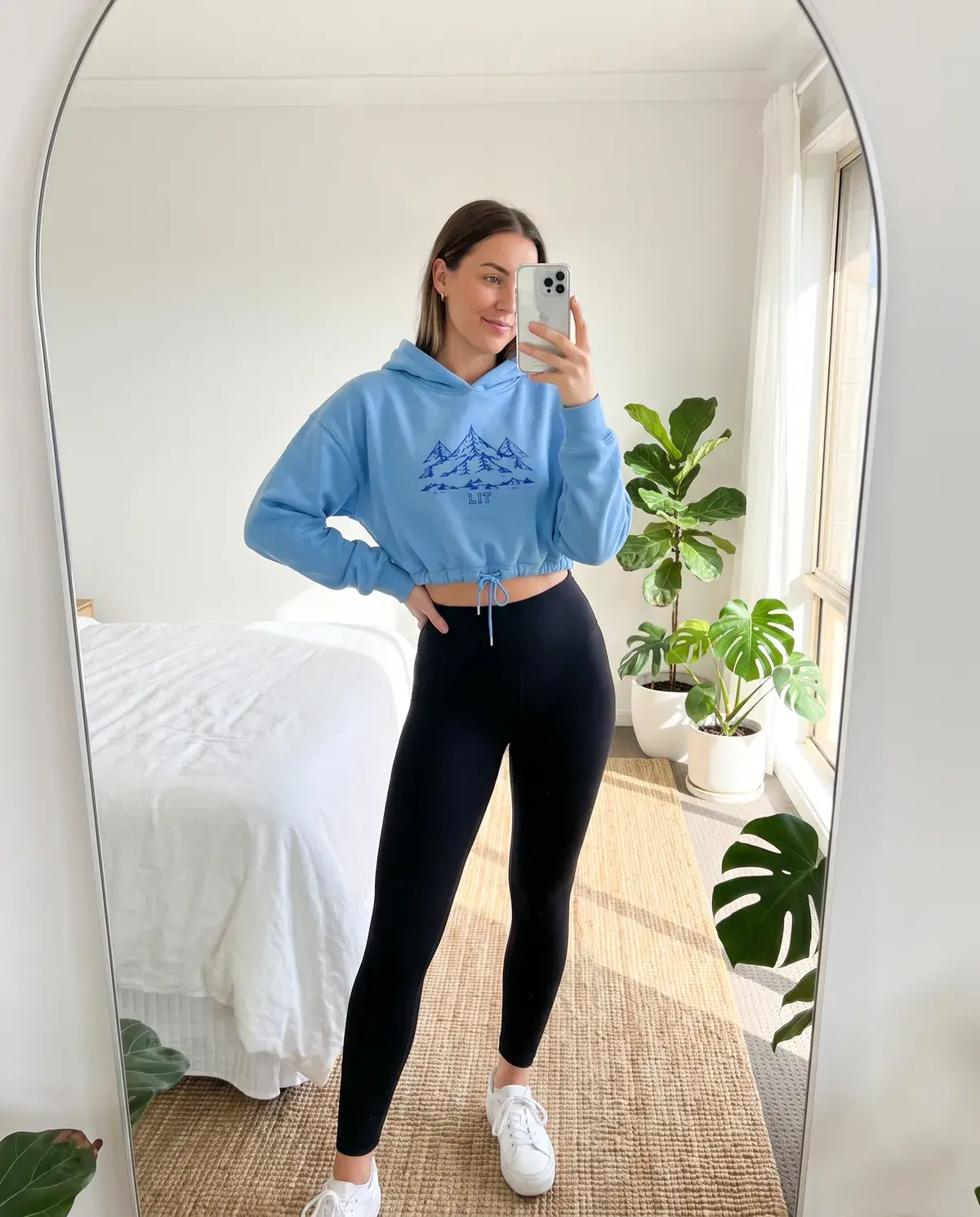 A woman takes a mirror selfie in a bright bedroom in the sky blue Cropped Mountain Hoodie and black leggings.
