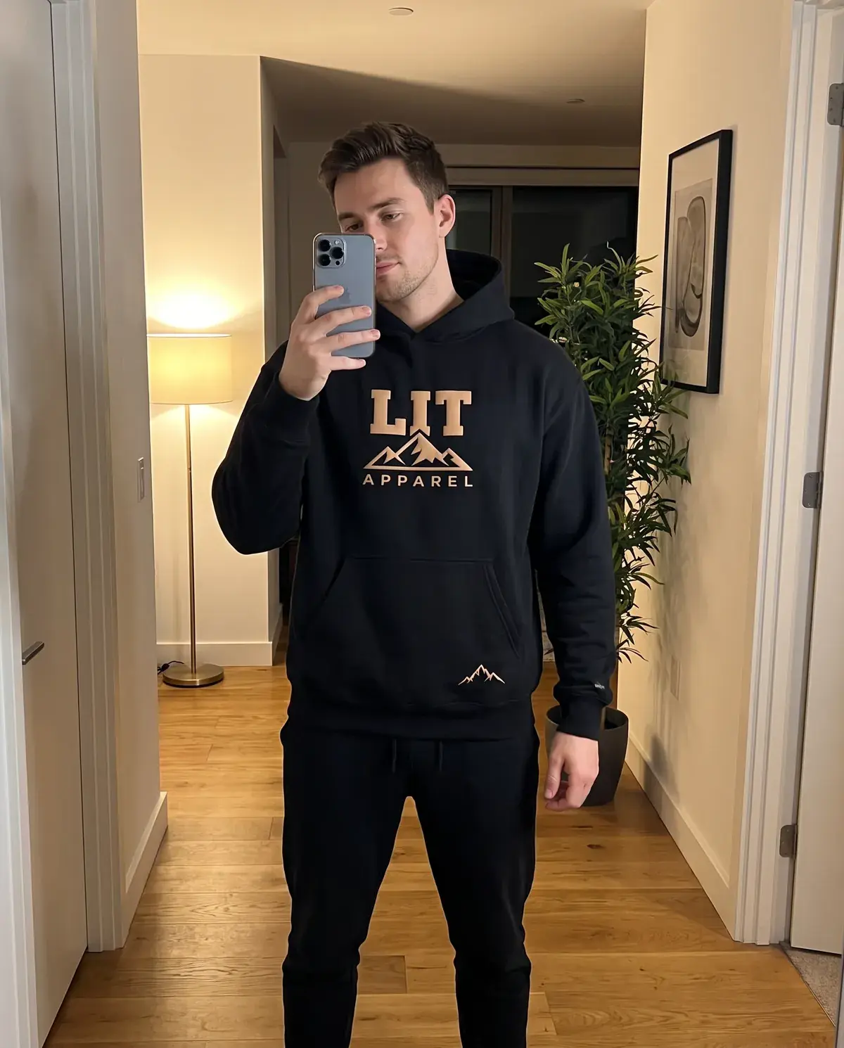 A man takes a mirror selfie in his hallway in the black LIT Logo Hoodie, its tan LIT mountain logo on the chest.