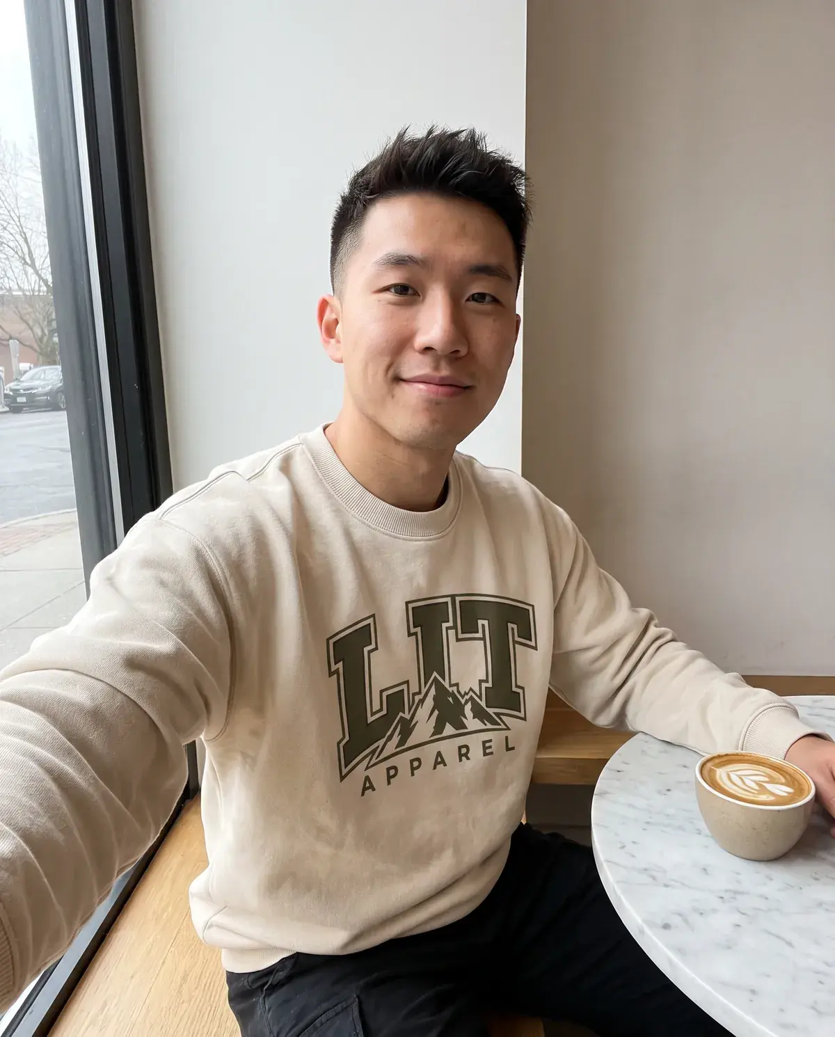 A man takes a selfie at a café window in the cream LIT Varsity Crewneck, a latte beside him.