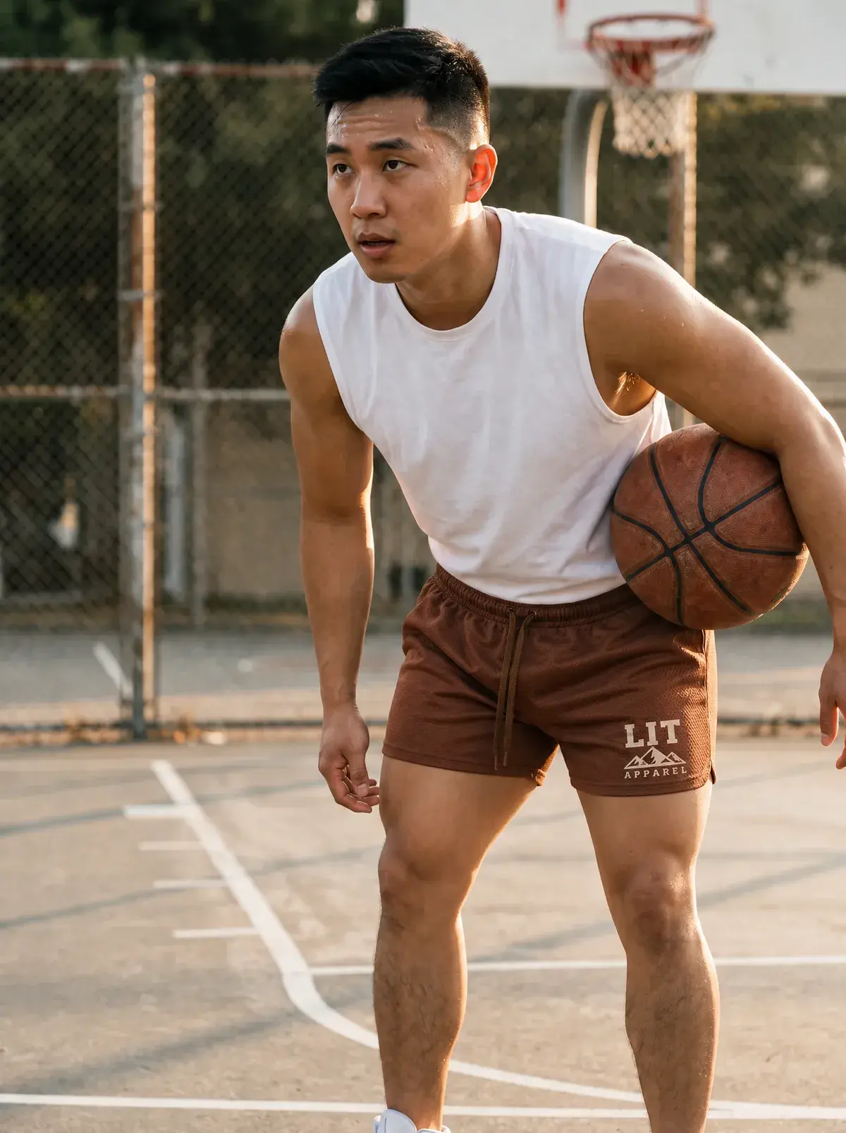 A man in a white tank and brown mesh shorts holds a basketball on an outdoor court.