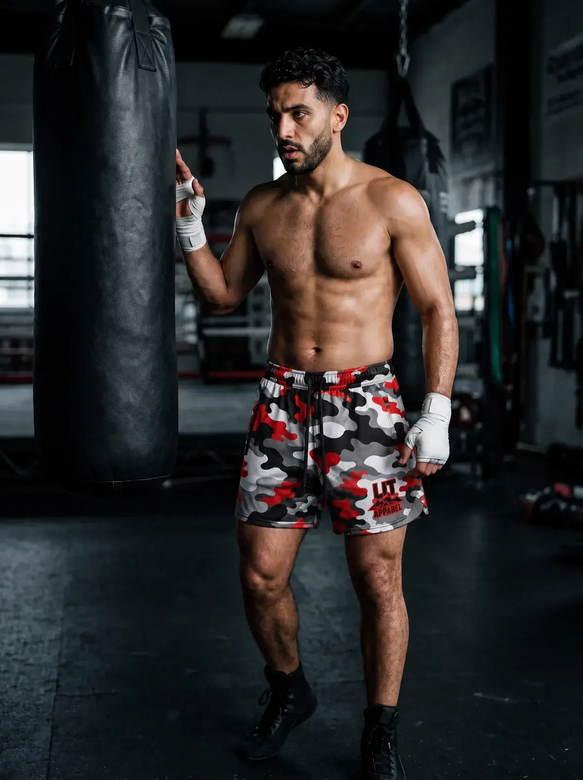 A boxer in hand wraps and red camo mesh shorts stands by a heavy bag in a dark gym.