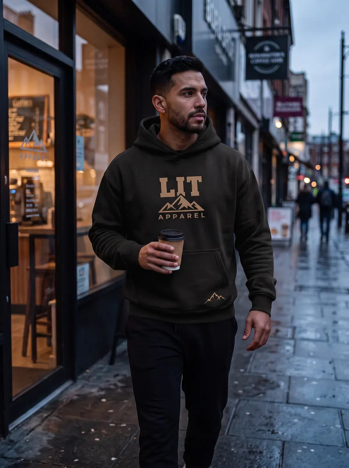A man in a black LIT hoodie walks out of a coffee shop at dusk, cup in hand.
